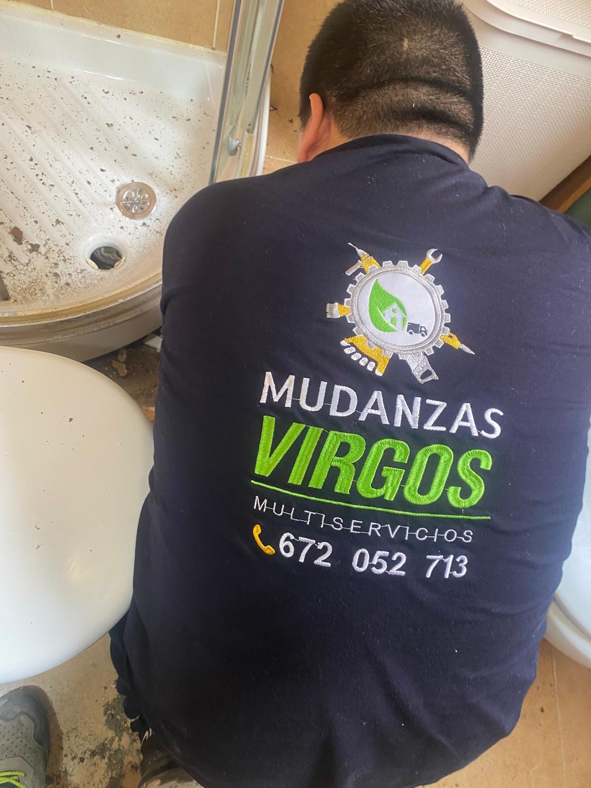 Fontanero de espaldas, con el logo de Mudanzas Virgos en la camiseta, revisando el desagüe bajo un plato de ducha
