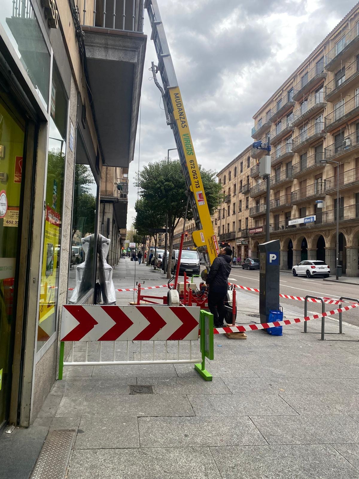 Grúa elevadora amarilla rotulada con Mudanzas Virgos montada en la acera, con la zona de trabajo vallada