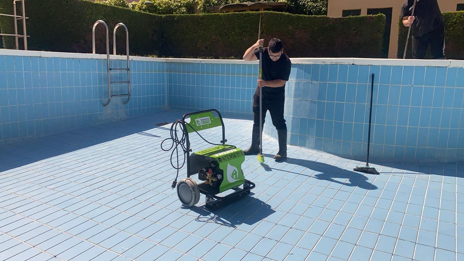 Operario limpiando el fondo de una piscina vacía de azulejo azul con una hidrolimpiadora verde