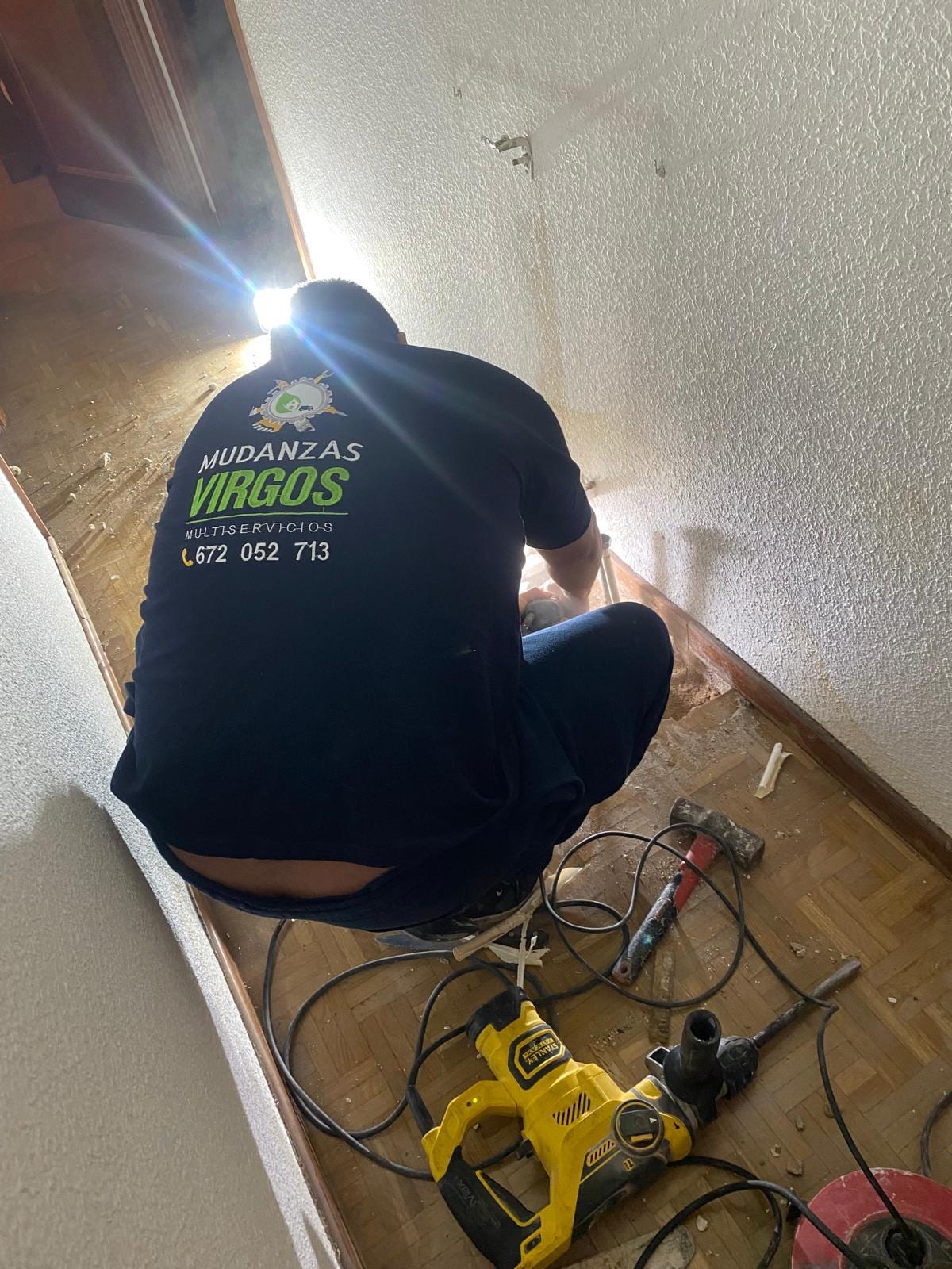 Técnico con camiseta de Mudanzas Virgos trabajando a ras de suelo en un pasillo, con taladro y herramientas junto a la pared abierta